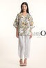 A woman wears the LinenO French Linen Blouse TP1714P-11 Bianco, featuring a loose fit and floral pattern, paired with white linen pants and open-toed sandals. She has long dark hair. The background is plain white with "LINEN ORIGINS.