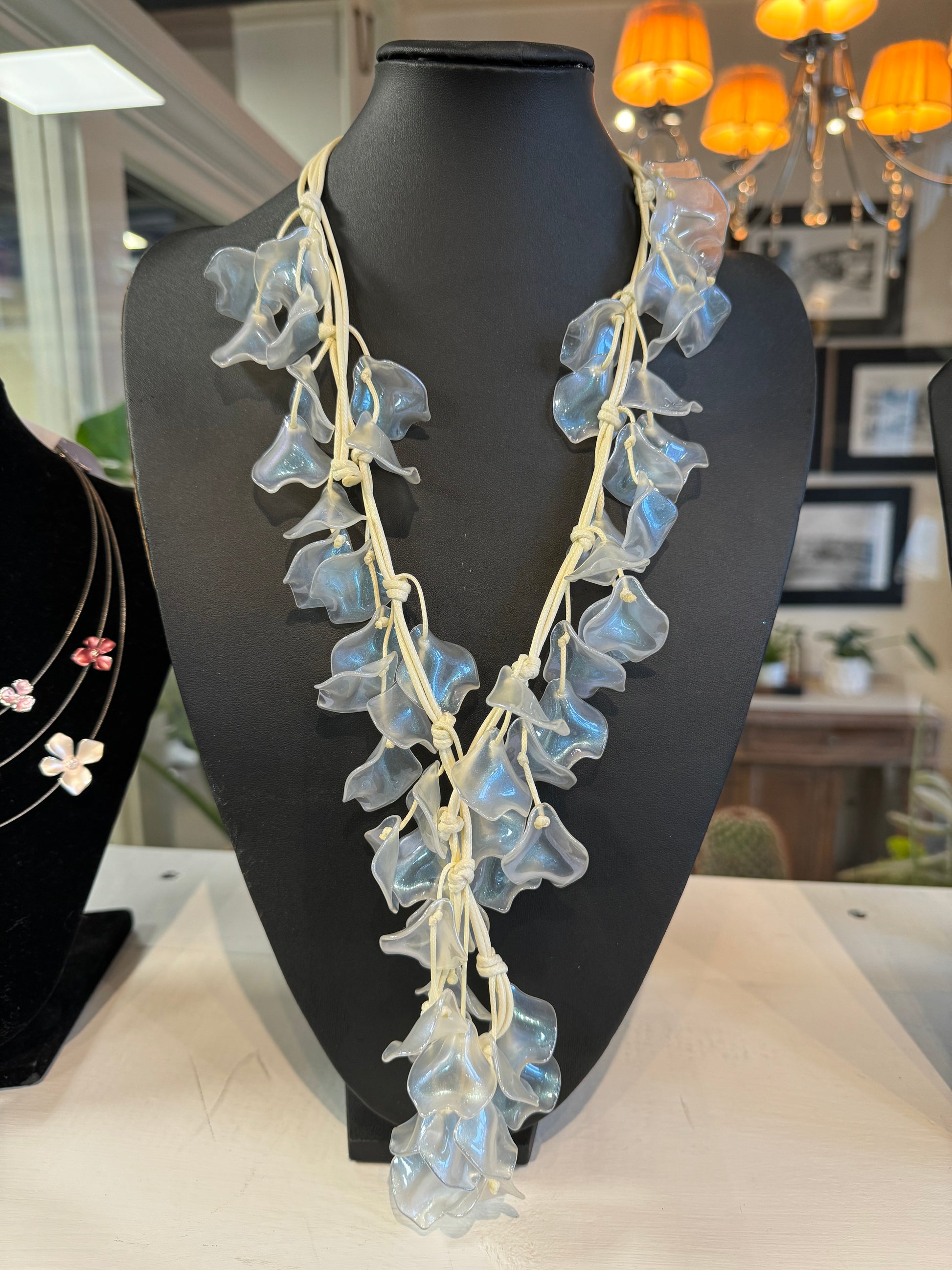 A stylish accessory, the Translucent Algae Necklace with Earrings by Posies Flowers & Fashion Inc., is displayed on a black bust. It features translucent blue glass flowers interwoven with beige cords. The backdrop has warm lighting, with blurred wall frames and chandeliers adding a cozy ambiance. Additional jewelry pieces are partially visible in the background.