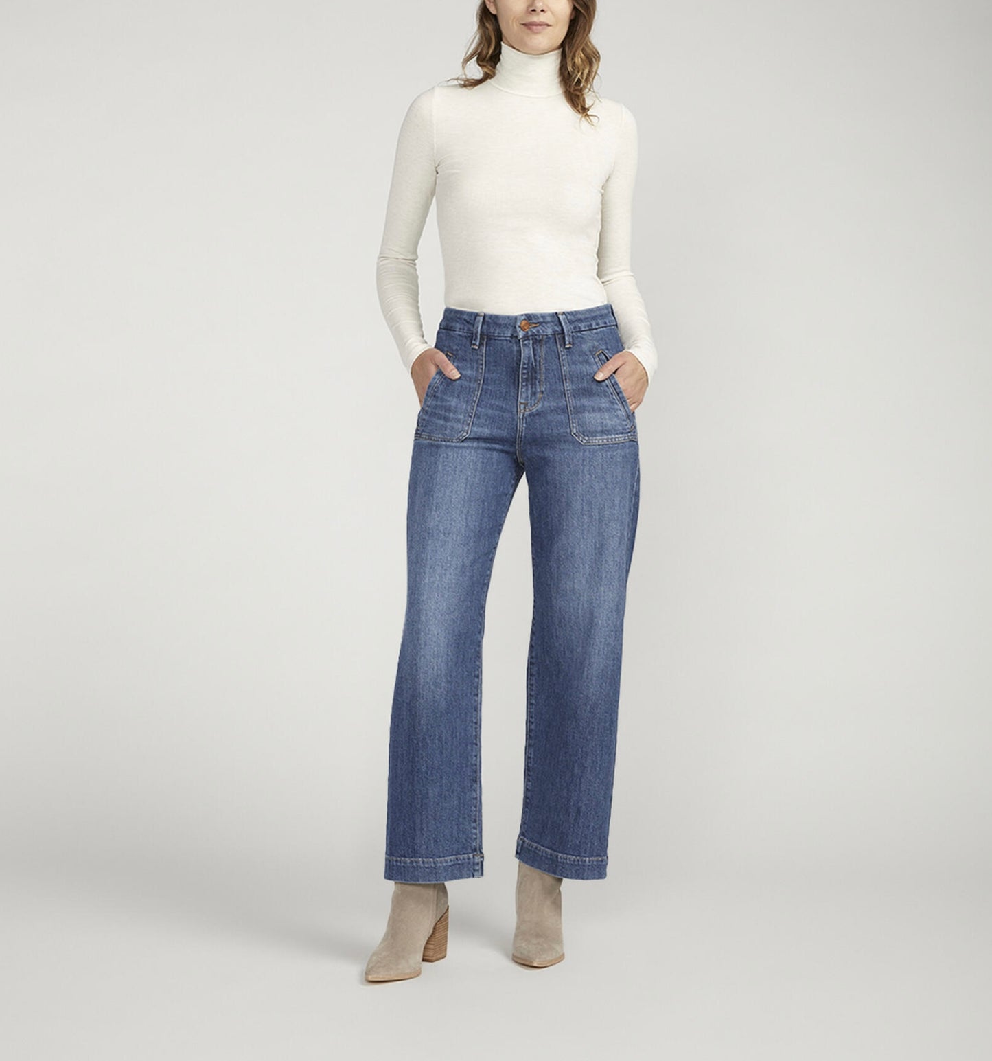 A person standing against a plain background is wearing a white turtleneck long-sleeve shirt, **JAG's Sophia High Rise Wide Leg Jeans in medium indigo wash with front pockets**, and light-colored ankle boots. Their hands are in their pockets and they are facing forward.