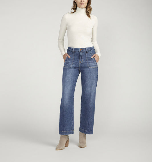 A person standing against a plain background is wearing a white turtleneck long-sleeve shirt, **JAG's Sophia High Rise Wide Leg Jeans in medium indigo wash with front pockets**, and light-colored ankle boots. Their hands are in their pockets and they are facing forward.