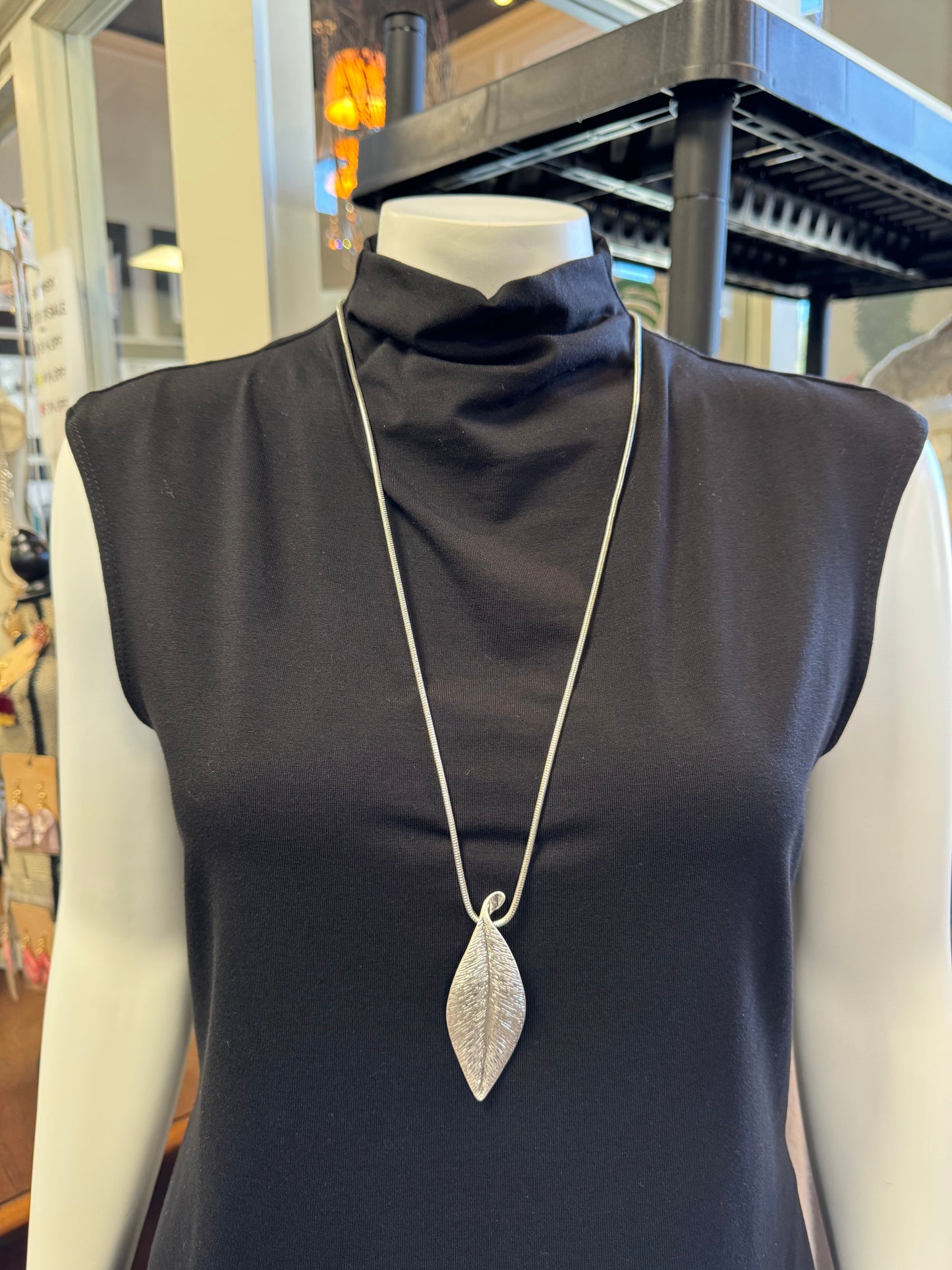 A mannequin displays a sleeveless black top, beautifully accentuated by the Andy 16 Necklace from Elise Accessories, which features a striking long silver chain with a large leaf-shaped pendant. In the background, shelves showcase an enticing jewelry collection alongside various store displays.