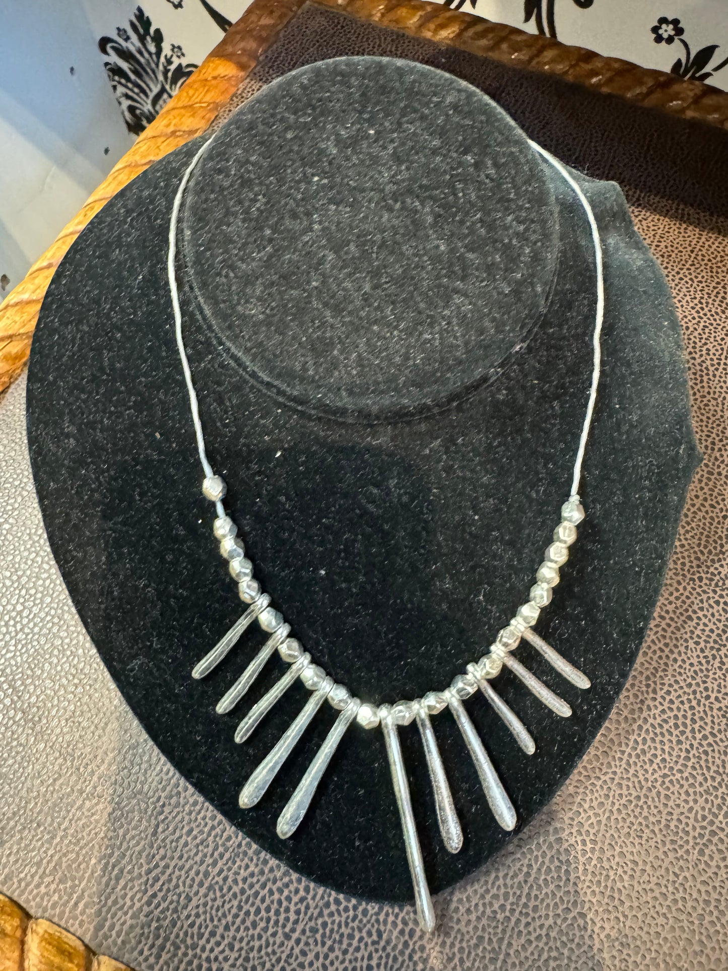 The Suzie Blue Silver Necklace is beautifully showcased on a black velvet jewelry display stand. This elegant piece features a combination of small round beads and eleven elongated teardrop-shaped beads hanging in a row, creating a sophisticated and stylish presentation with its simple design.