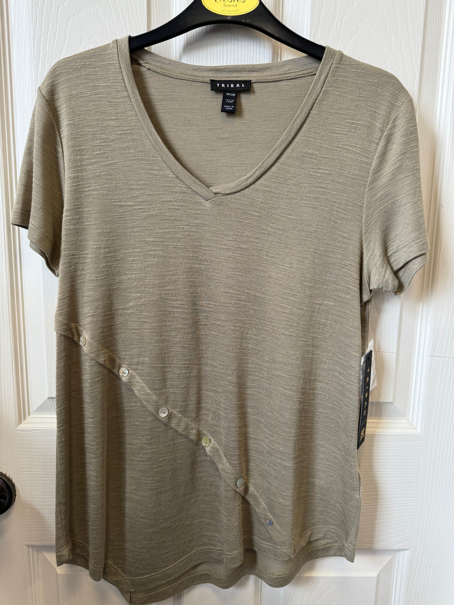 A moss green T-shirt from the brand Tribal hangs on a black hanger against a white wooden door. The shirt, part of a stylish piece, has a plain design with a soft texture. A small tag is visible on the left side, near the bottom. The doorknob on the left side of the image is black.