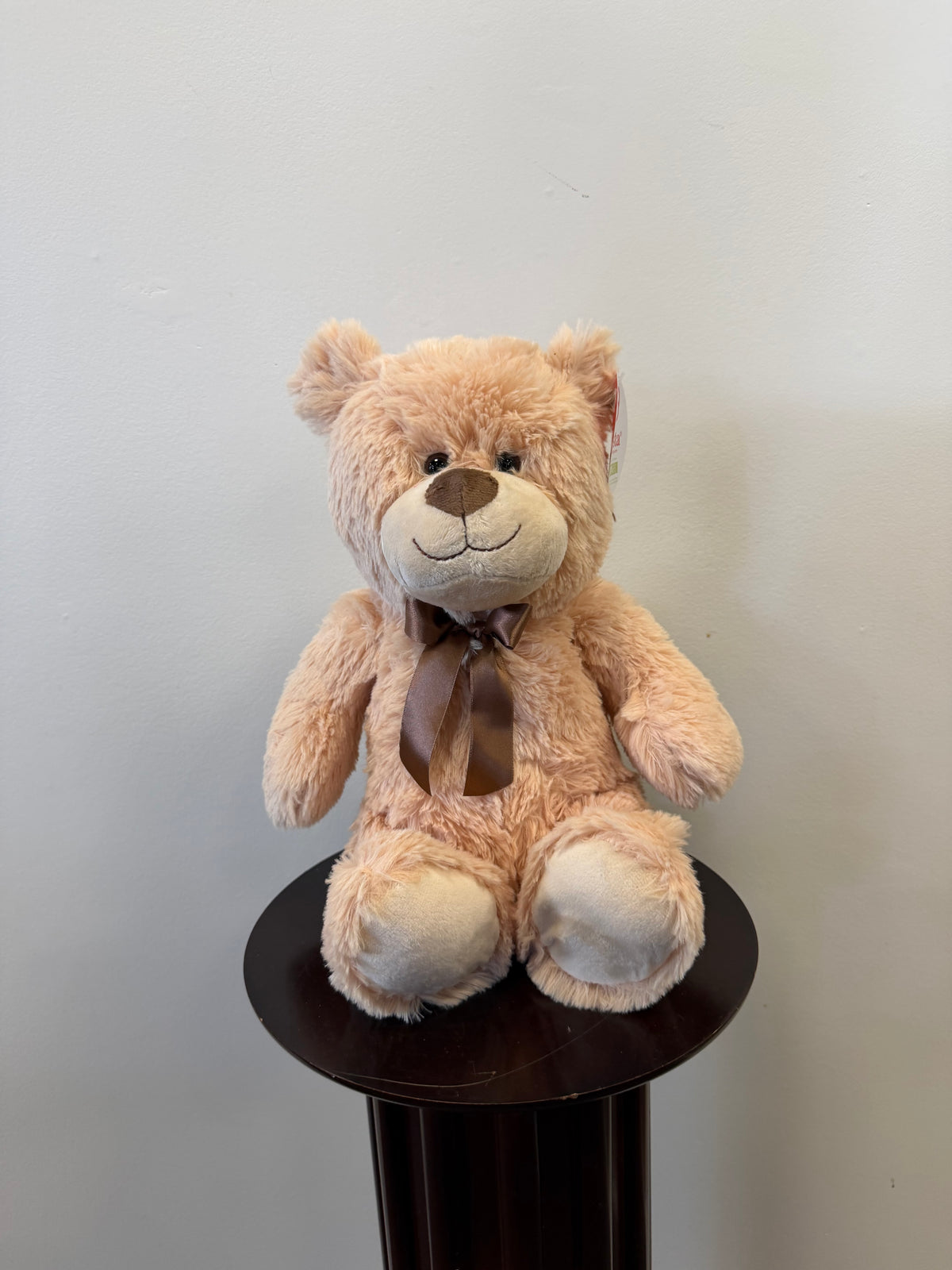 The Fiesta Classic Teddy Bear - Tan 13 inch, featuring a dark brown ribbon, is elegantly displayed on a round, dark wooden pedestal against a plain light-colored wall.