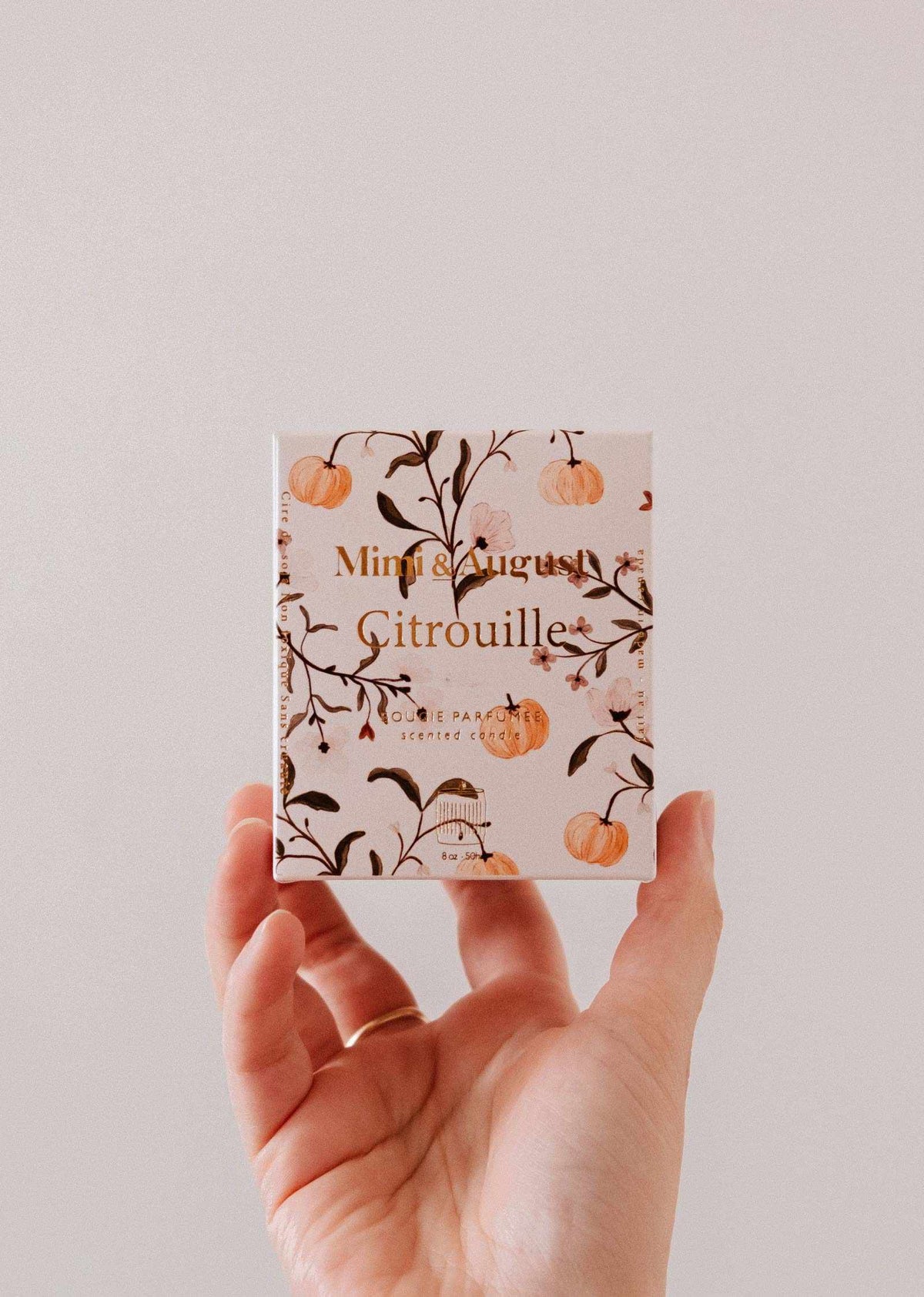 A hand holds a small square box adorned with orange pumpkins and vines. The box, containing the Citrouille - 8 oz reusable candle from Mimi & August, hints at its inviting autumn-inspired scent, all set against a plain light background.