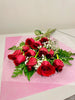 A dozen fresh-cut red roses from Posies Flowers & Fashion, enhanced with green fern leaves and baby's breath, are beautifully arranged on pink wrapping paper against a light-colored background.