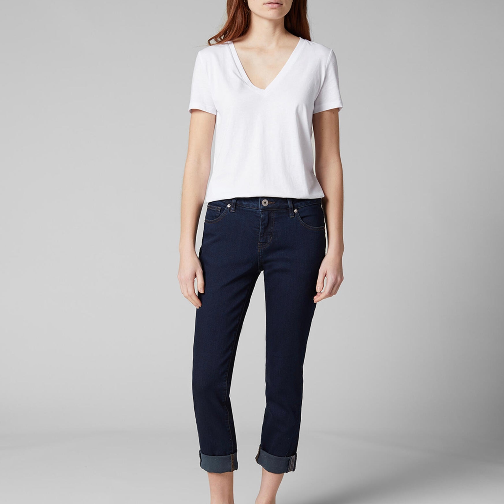 A person stands against a plain grey background wearing a white V-neck T-shirt, dark blue Carter Mid Rise Girlfriend Jeans by JAG made from Sustainable Premium Stretch denim with rolled cuffs, and has hands resting at sides. The person has long, straight hair.