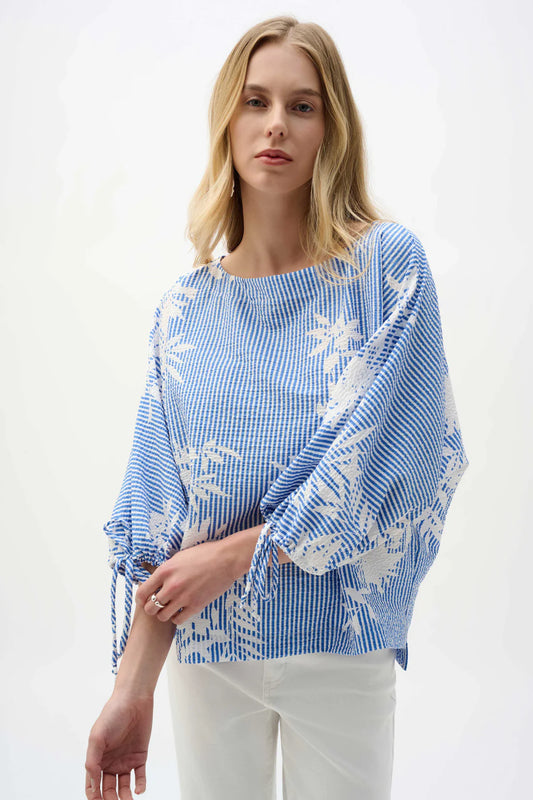 A woman with blonde hair models the Joseph Ribkoff Seersucker Blouse 262067, featuring a loose blue and white pattern with dolman sleeves, paired with white pants against a plain white background.