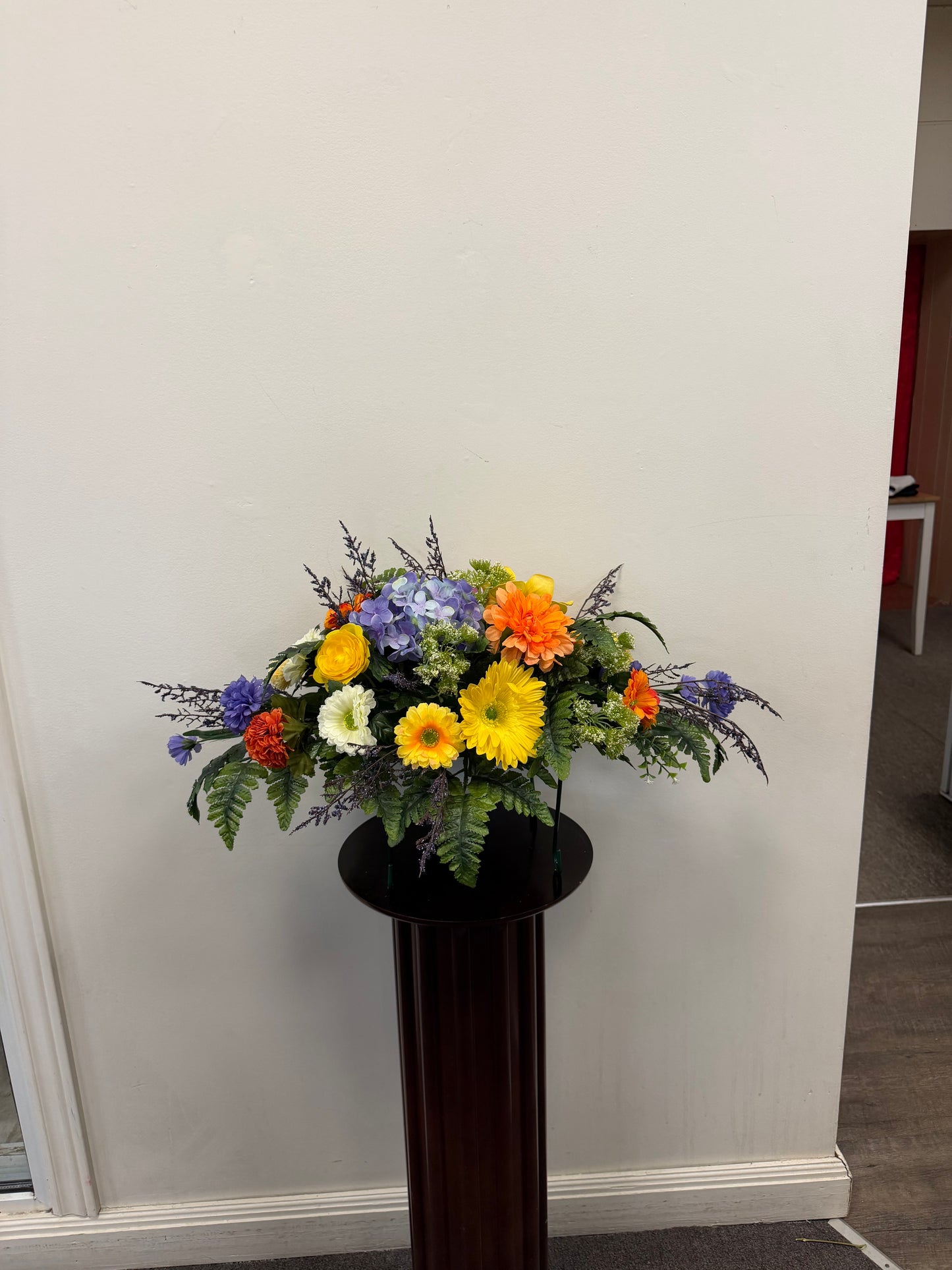 The 11 - Tombstone Saddle (60 cm) by Posies Flowers & Fashion Inc. features vibrant yellow, orange, white, and purple flowers with green foliage, displayed on a tall dark stand against a plain white wall.