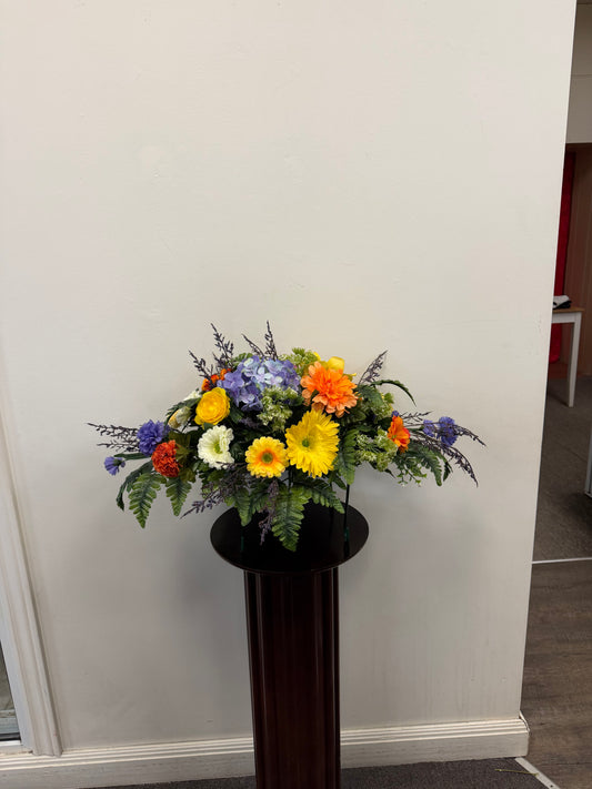 The 11 - Tombstone Saddle (60 cm) by Posies Flowers & Fashion Inc. features vibrant yellow, orange, white, and purple flowers with green foliage, displayed on a tall dark stand against a plain white wall.