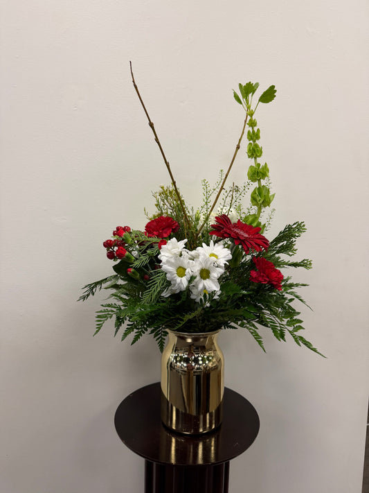 The Sign of the Times Christmas Vase by Posies Flowers & Fashion features a gold cylinder vase on a dark stand, filled with red and white flowers, lush greenery, and tall branches, set against a plain white background.