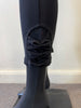 A close-up of a sleek black mannequin leg wearing Bali Corp's Pant 8622, crafted from polyester and spandex. These elegant leggings feature decorative bows on the lower cuff, enhancing the sleek design. The mannequin stands on a square base against a pale wall.