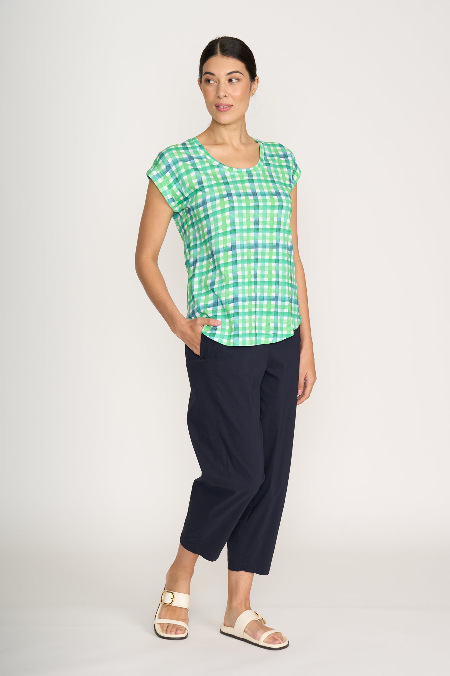A woman wears the FOIL Role Model Tee Edgy FO8173 in green and blue plaid, paired with dark navy cropped pants and white sandals. She stands against a plain background, one hand in her pocket, looking to the side.