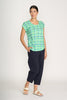 A woman wears the FOIL Role Model Tee Edgy FO8173 in green and blue plaid, paired with dark navy cropped pants and white sandals. She stands against a plain background, one hand in her pocket, looking to the side.