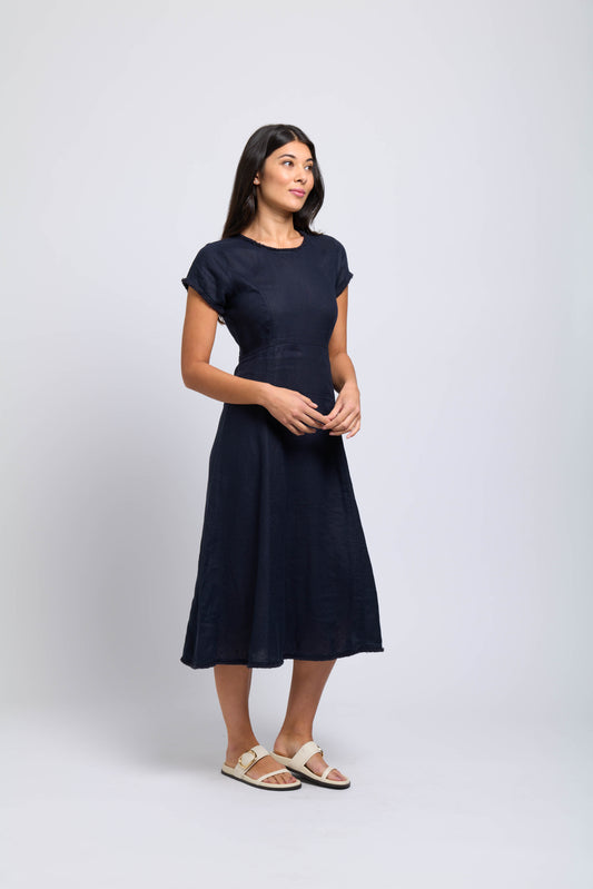 A woman with long dark hair wears the FOIL Fringe Event Dress FO7890 in Midnight and light sandals, standing against a plain light background, looking to the side with a soft smile.