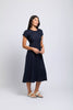 A woman with long dark hair wears the FOIL Fringe Event Dress FO7890 in Midnight and light sandals, standing against a plain light background, looking to the side with a soft smile.