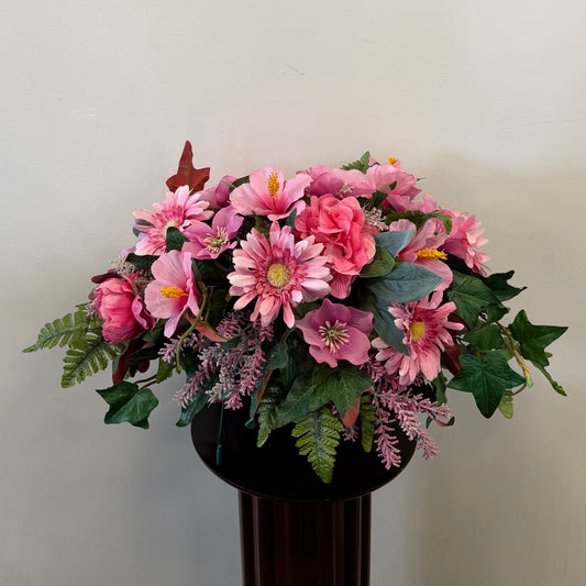The 9 - Tombstone Saddle 60 cm Length by Posies Flowers & Fashion Inc. features pink flowers, green leaves, and ferns in a black vase—ideal as a silk cemetery flower arrangement or tombstone saddle.