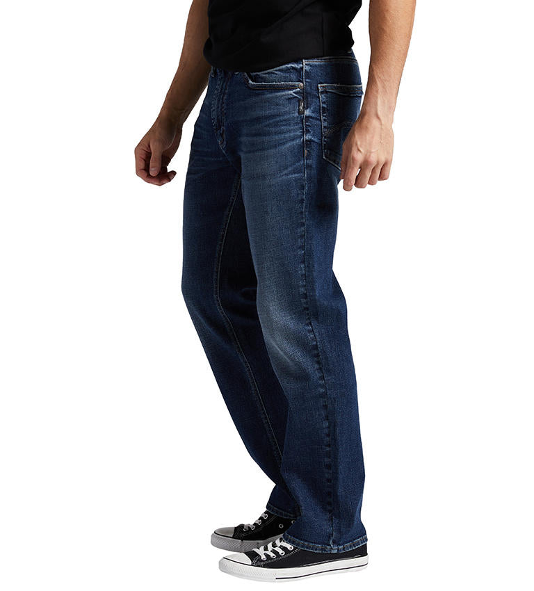 A person is standing, wearing Silver Jeans Co.'s Grayson Classic Fit Straight Leg Jeans in a dark indigo wash with classic five-pocket styling. The slightly faded straight-leg jeans are paired with a black shirt and black low-top sneakers with white soles. The background is white.