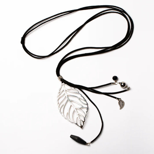 The Suzie Blue Long Leaf Pendant On Suede With Charms in black features a large silver leafy pendant, adorned with small decorative charms including a handmade silver leaf, beads, and a black stone. The ethically sourced necklace is laid out on a white background.