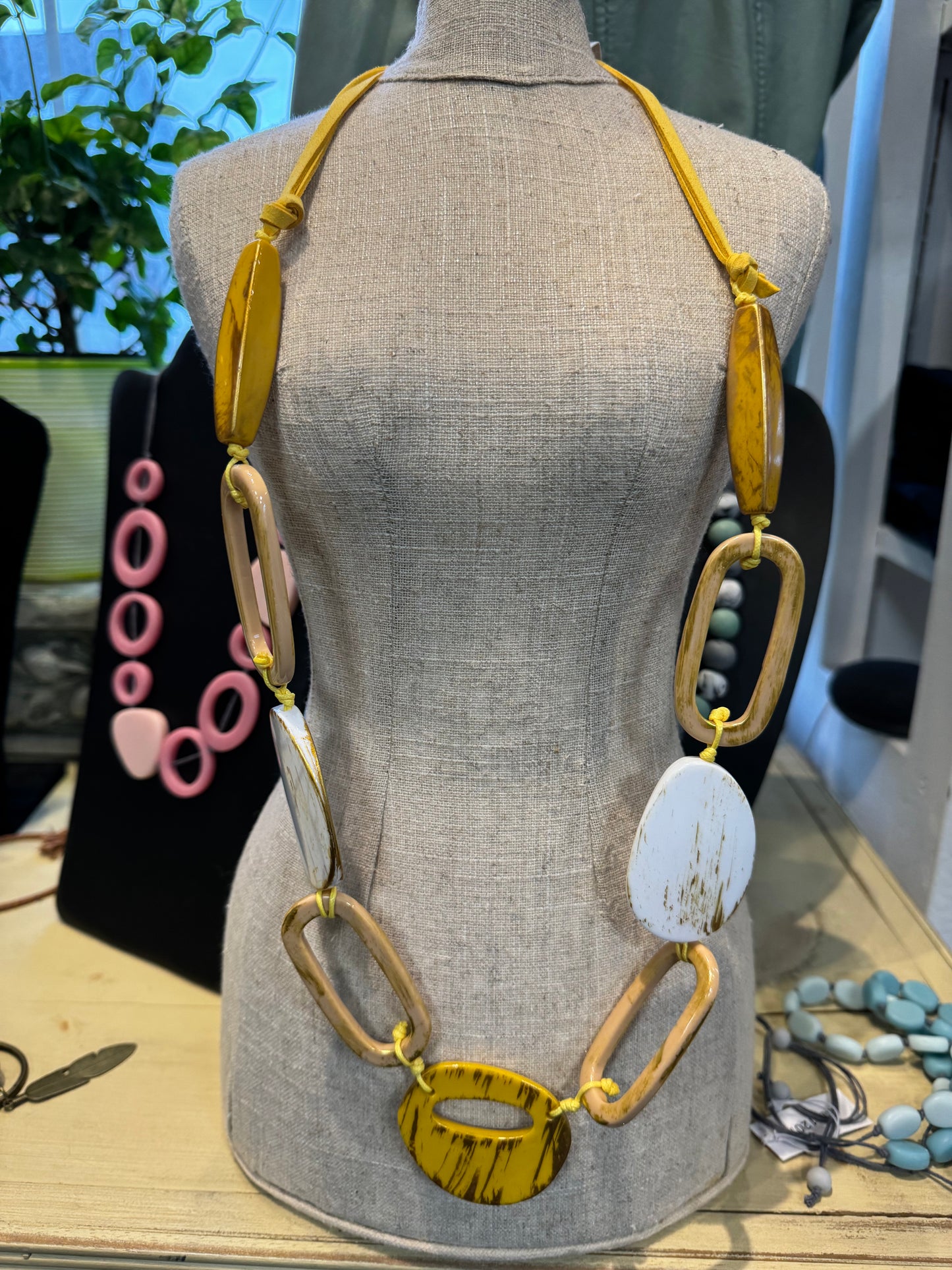 A beige dress form showcases a stylish ensemble featuring the Yellow and White Coloured Loop Necklace With Earrings by Posies Flowers & Fashion Inc. The necklace is adorned with large, chunky geometric beads in various shades of yellow, brown, and white. In the background, there is a green plant along with earrings and other jewelry pieces, including a necklace with pink beads displayed on a black stand.