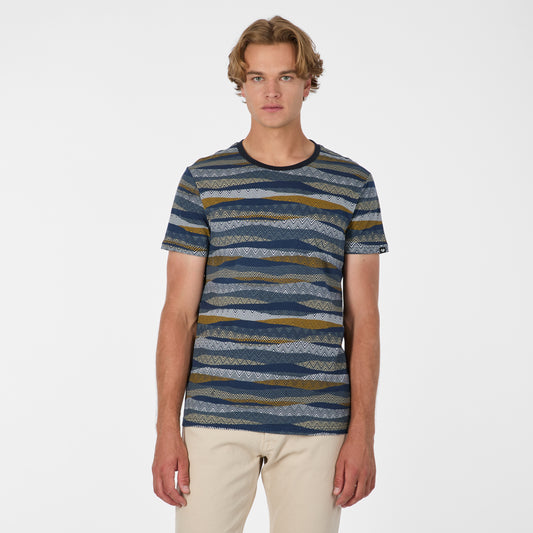 A young man with wavy blond hair, wearing RAGWEAR's T-shirt Wannel 2612-15050 Grey Multi-Coloured and light beige pants, stands facing forward against a plain white background.