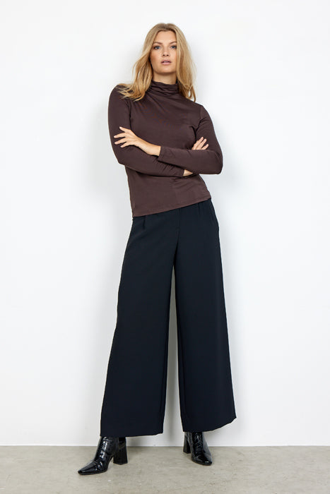 A person stands against a plain white background, arms crossed, wearing a dark brown Soyaconcept SC-MARICA 12 t-shirt, wide-leg black pants, and black ankle boots. Their expression appears neutral, and their body is slightly leaning to one side.