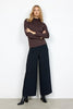 A person stands against a plain white background, arms crossed, wearing a dark brown Soyaconcept SC-MARICA 12 t-shirt, wide-leg black pants, and black ankle boots. Their expression appears neutral, and their body is slightly leaning to one side.