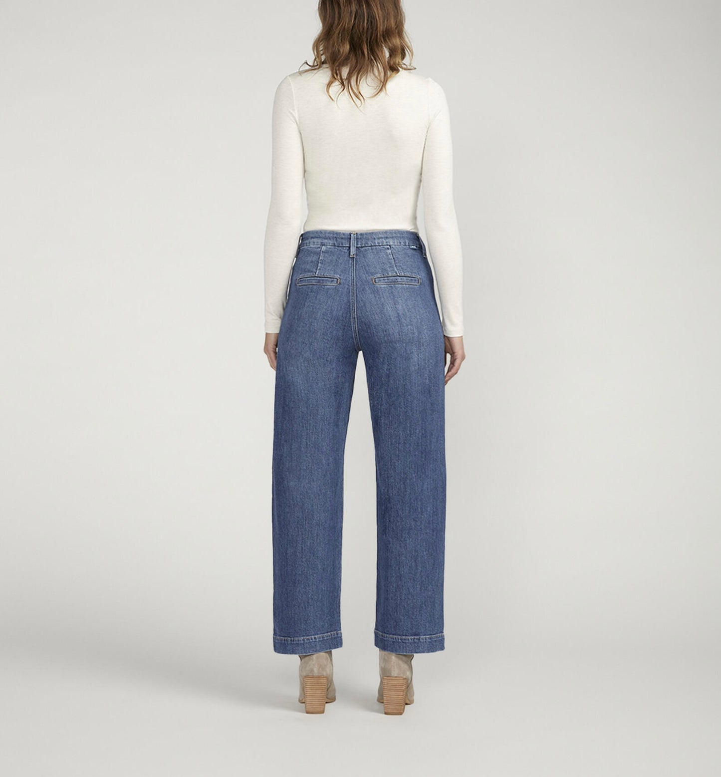 A person standing against a plain background is wearing a white turtleneck long-sleeve shirt, **JAG's Sophia High Rise Wide Leg Jeans in medium indigo wash with front pockets**, and light-colored ankle boots. Their hands are in their pockets and they are facing forward.