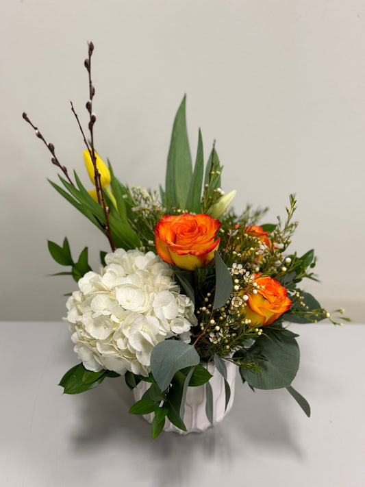 Introducing "Orange Delight" by Posies Flowers & Fashion: a striking floral arrangement set in a pristine white vase, showcasing vibrant orange roses, elegant white hydrangeas, petite yellow blossoms, lush green foliage, and delicate white sprigs. Please note that substitutions may occur based on availability.