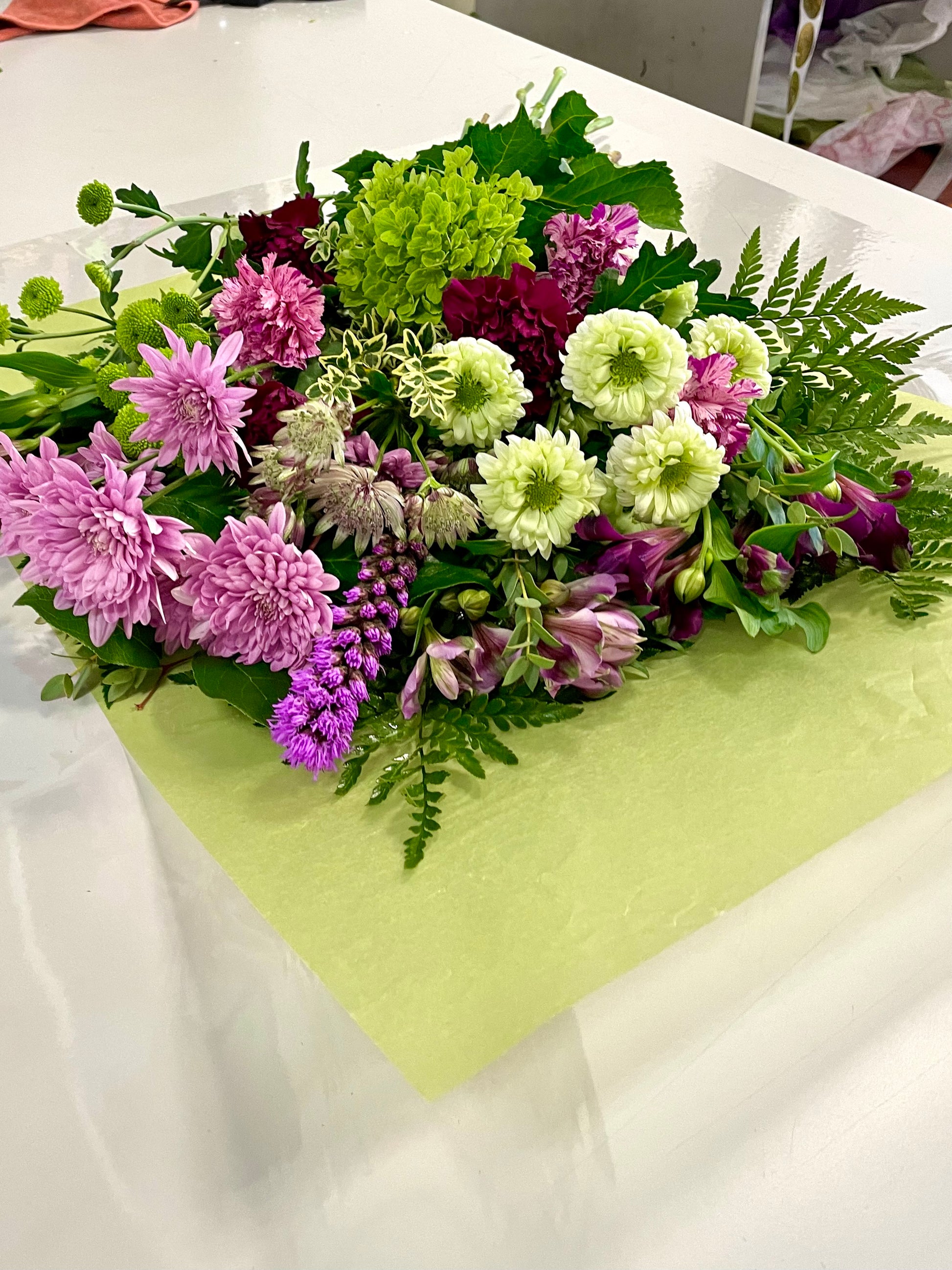 A stunning bouquet called Designer’s Choice Freshcuts from Posies Flowers & Fashion is resting on a white surface. The arrangement features vibrant orange and pink roses, cheerful yellow chrysanthemums, elegant white flowers, and assorted green foliage. It is partially wrapped in brown paper and elegantly tied with a burgundy ribbon.