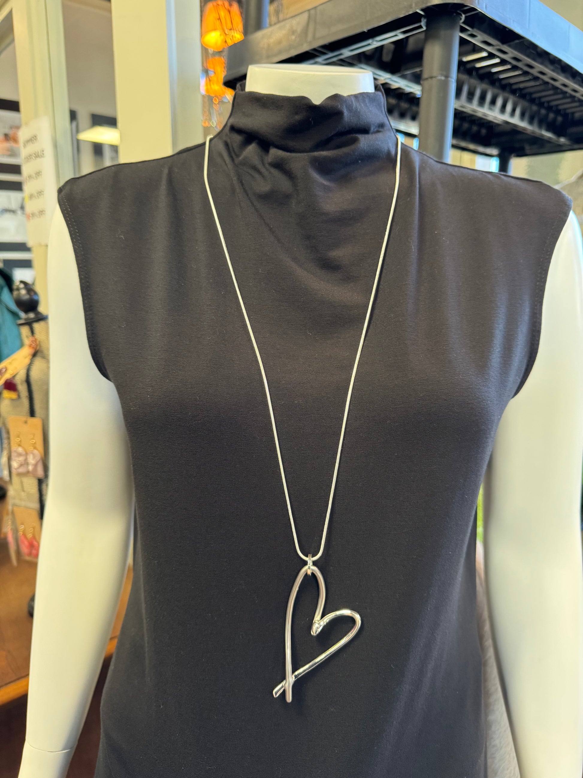 Showcased on a mannequin wearing a sleeveless black turtleneck top is the chic fashion piece, Necklace 7 by Elise Accessories—a long silver necklace featuring a stylized heart pendant. The background highlights shelves and items from an exclusive jewelry collection.