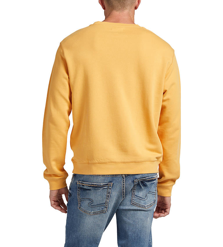 A man wearing a "HONEY" French terry cotton men's crewneck sweatshirt by Silver Jeans Co., featuring "49.8951 N -97.1384 W" embroidered on the front, paired with blue jeans. The sweatshirt includes ribbed cuffs and a crew neckline. He is looking slightly to the side, with one hand by his side and the other partially in a pocket.