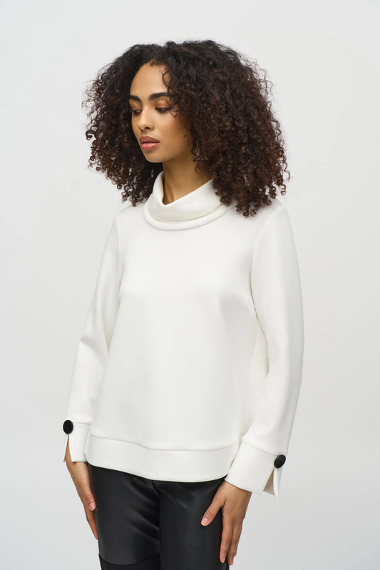 A woman with curly hair wears a loose-fitting, long-sleeved white Scuba Jersey Boxy Top 244129 by Joseph Ribkoff. The top, featuring button details on the sleeves, lends a boxy silhouette. She stands in front of a plain background, looking slightly downward with a neutral expression.