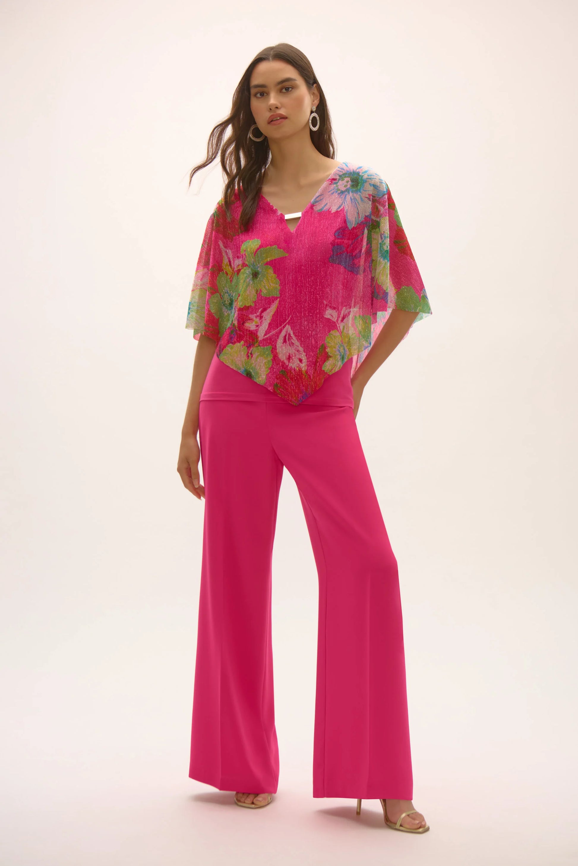 Wearing Joseph Ribkoff's Signature Metallic Pleated Knit Floral Print Poncho Top, paired with bright pink wide-legged pants, a person casually tucks one hand in a pocket, striking a confident pose against a plain background.