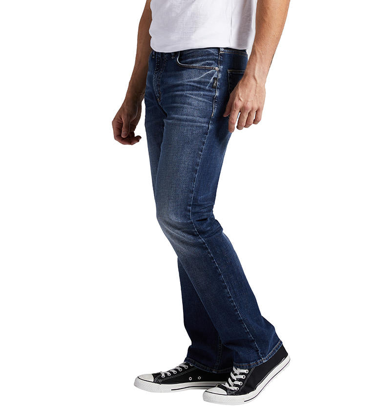 A person stands with arms relaxed at their sides, wearing a white t-shirt, Silver Jeans Co.'s Machray Athletic Fit Straight Leg Jeans in dark blue, and black sneakers with white soles. The person is shown from the shoulder down, without their face visible.