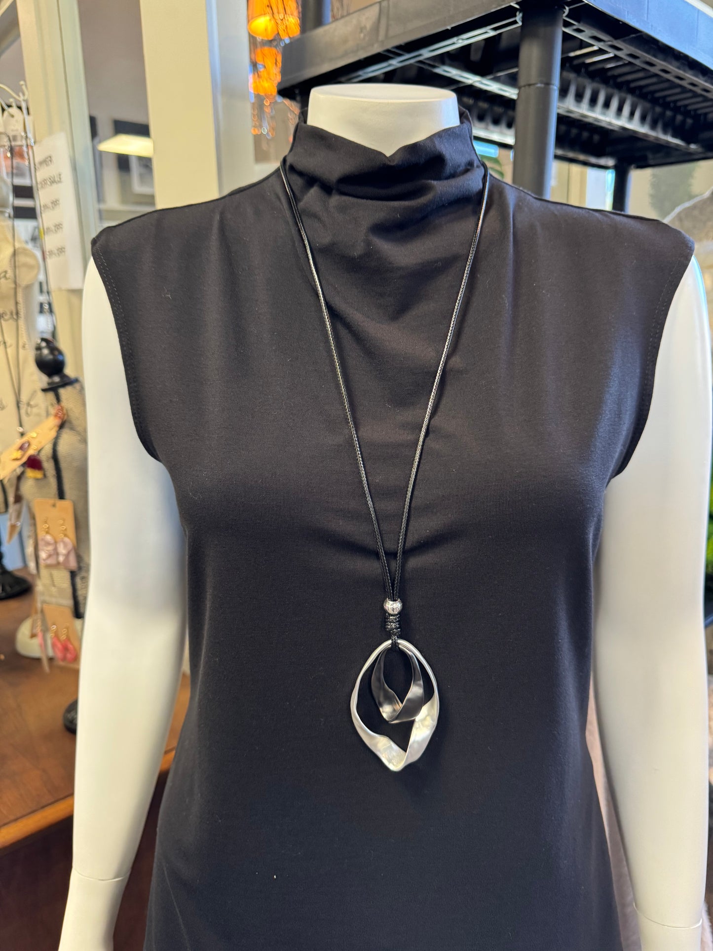 A mannequin showcases a sleeveless black turtleneck dress accented with the Sofia 11 Necklace from Elise Accessories, exuding elegance. The background includes various store items and decor.