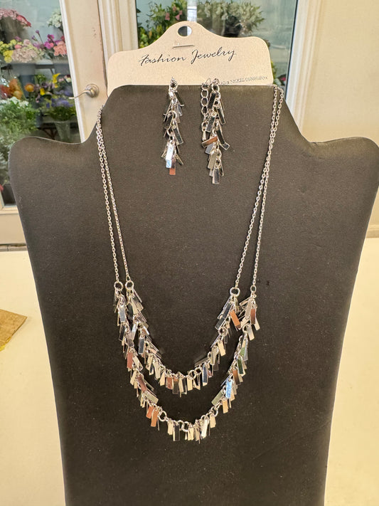 A display showcasing the chic "Flashy Necklace and Earrings" set by Posies Flowers & Fashion Inc., featuring a striking silver-toned necklace with two layers of dangling reflective charms, perfectly matched with earrings that share the same charm design. This elegant set is beautifully presented on a black stand.