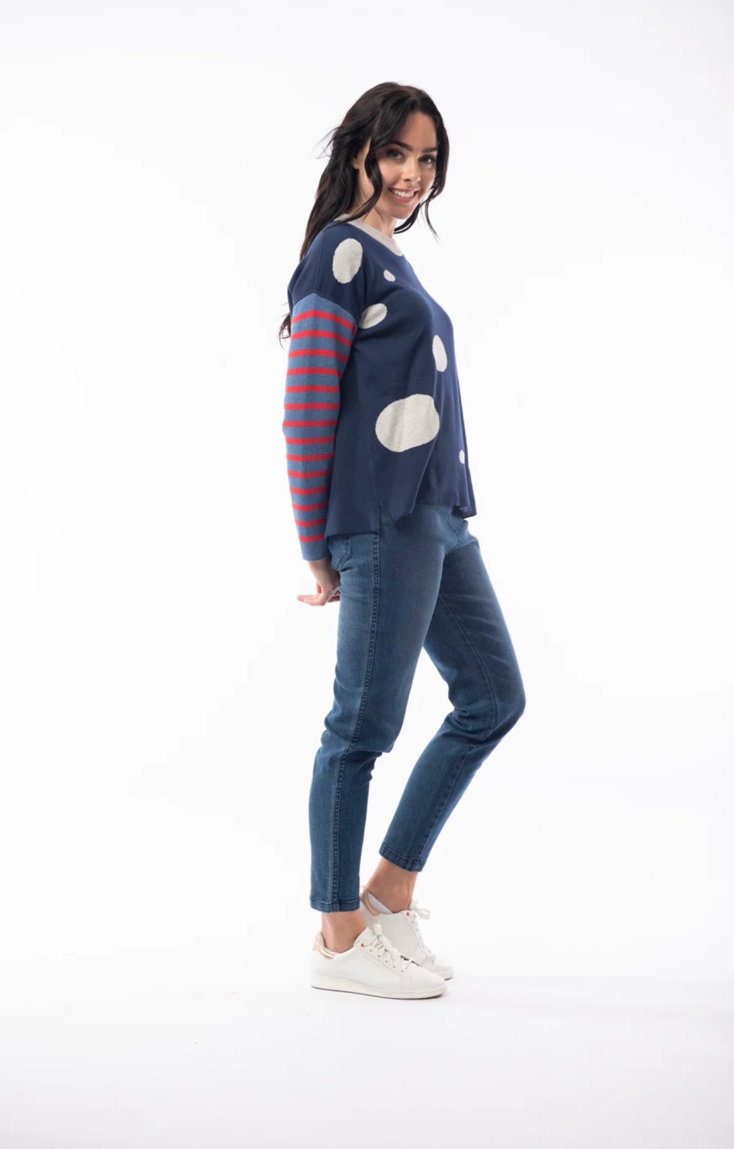 A woman with long dark hair is wearing the ORIENTIQUE - REVERSIBLE ROUND NECK SPOT/STRIPE JUMPER (1229) made of organic cotton, featuring a navy blue background adorned with white polka dots and red striped sleeves. She's paired it with blue jeans and white sneakers, and stands in a casual pose, slightly leaning back with her hands behind her. The background is plain white.