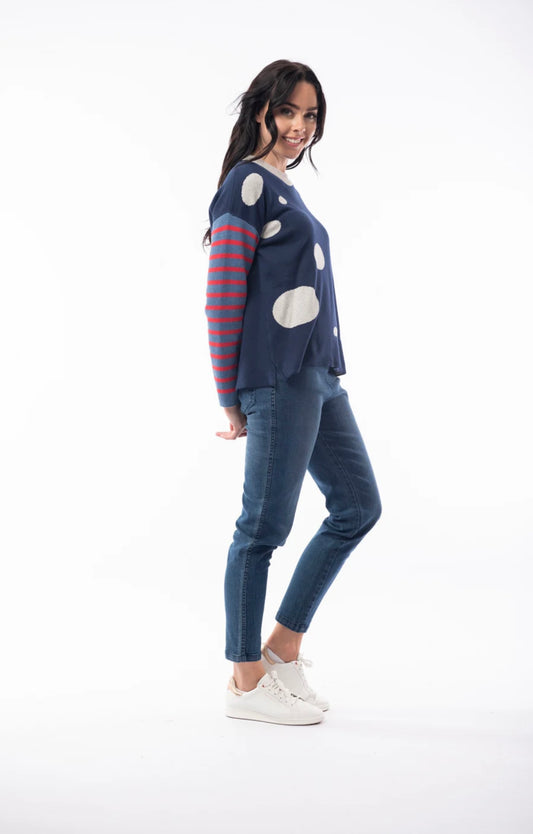 A woman with long dark hair is wearing the ORIENTIQUE - REVERSIBLE ROUND NECK SPOT/STRIPE JUMPER (1229) made of organic cotton, featuring a navy blue background adorned with white polka dots and red striped sleeves. She's paired it with blue jeans and white sneakers, and stands in a casual pose, slightly leaning back with her hands behind her. The background is plain white.