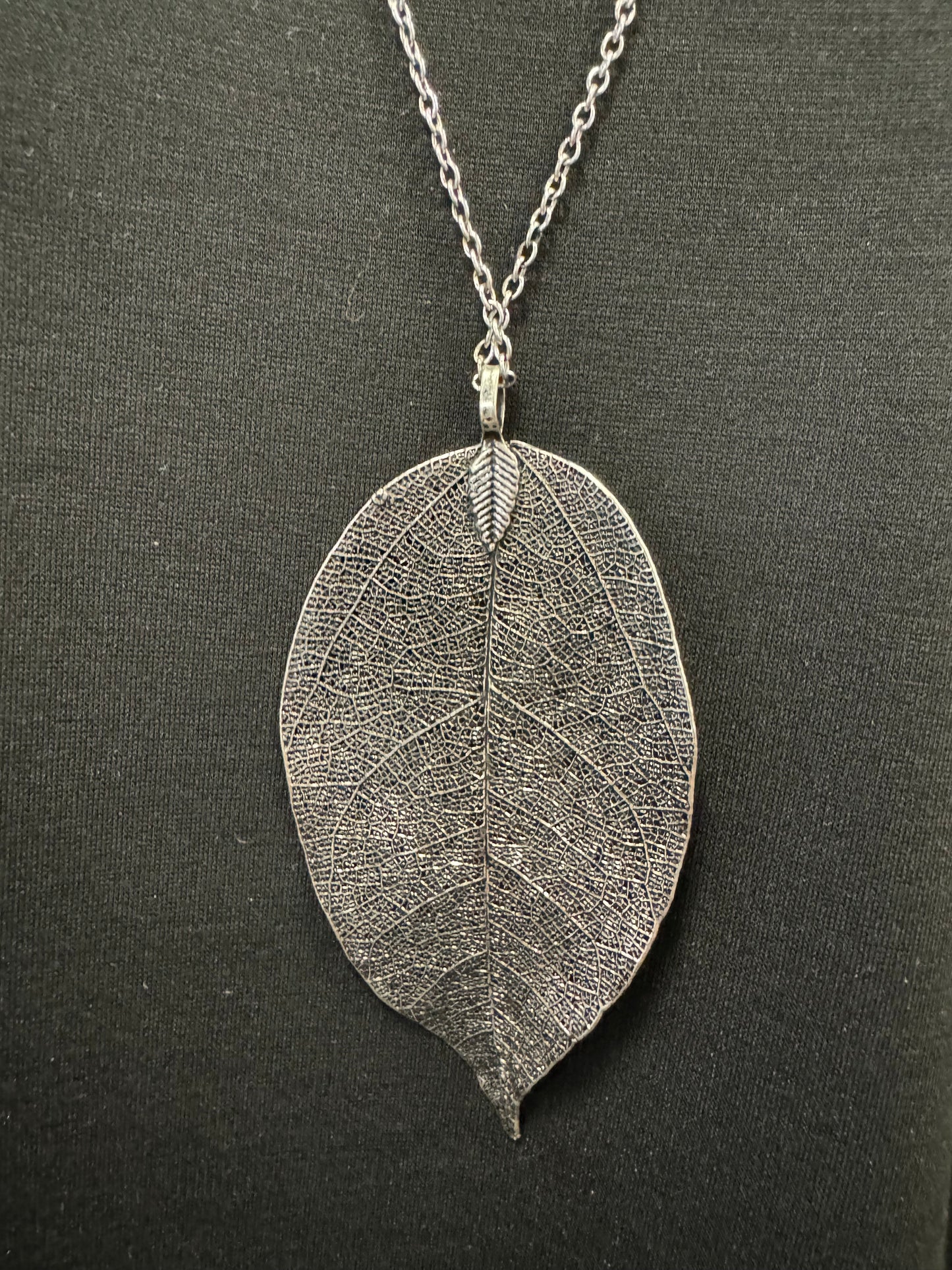 A black mannequin torso wears a black short-sleeve t-shirt and a long silver-colored chain featuring Posies Flowers & Fashion Inc.'s Dark Grey Coloured Leaf Pendant Costume Necklace. The background is plain and light-colored.