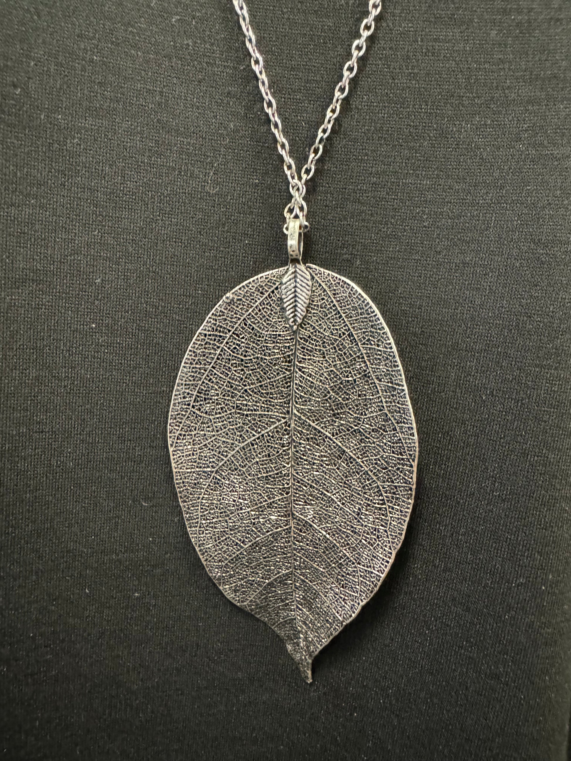 A black mannequin torso wears a black short-sleeve t-shirt and a long silver-colored chain featuring Posies Flowers & Fashion Inc.'s Dark Grey Coloured Leaf Pendant Costume Necklace. The background is plain and light-colored.