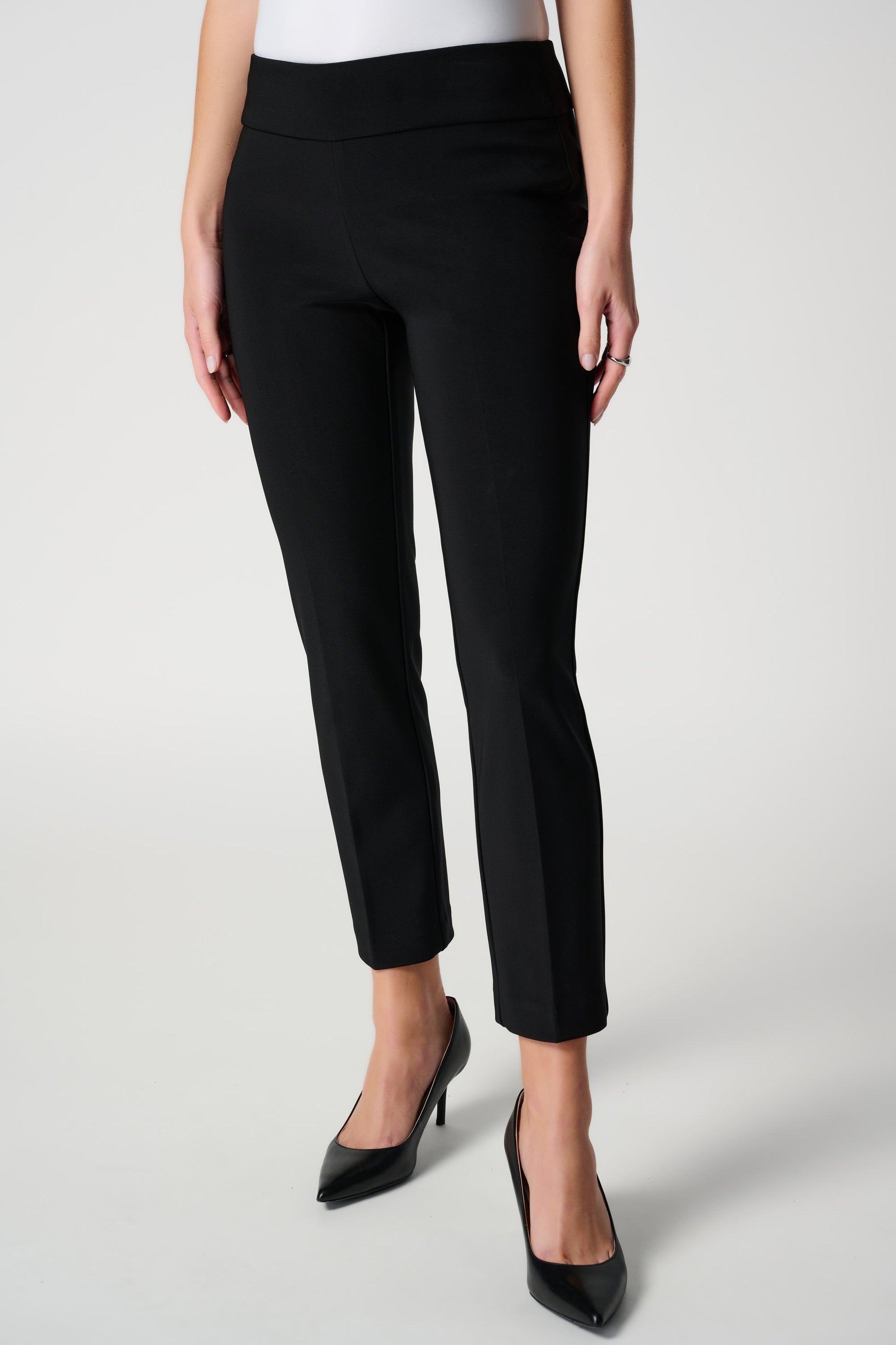 The individual is dressed in Joseph Ribkoff's Classic Cropped Pant 181089NOS, a pair of slim-fit black ankle pants, and black pointed-toe heels, standing against a plain light background. The tailored pants feature a simple, classic design with a smooth fit. They complement the white top that is partially visible in the image.