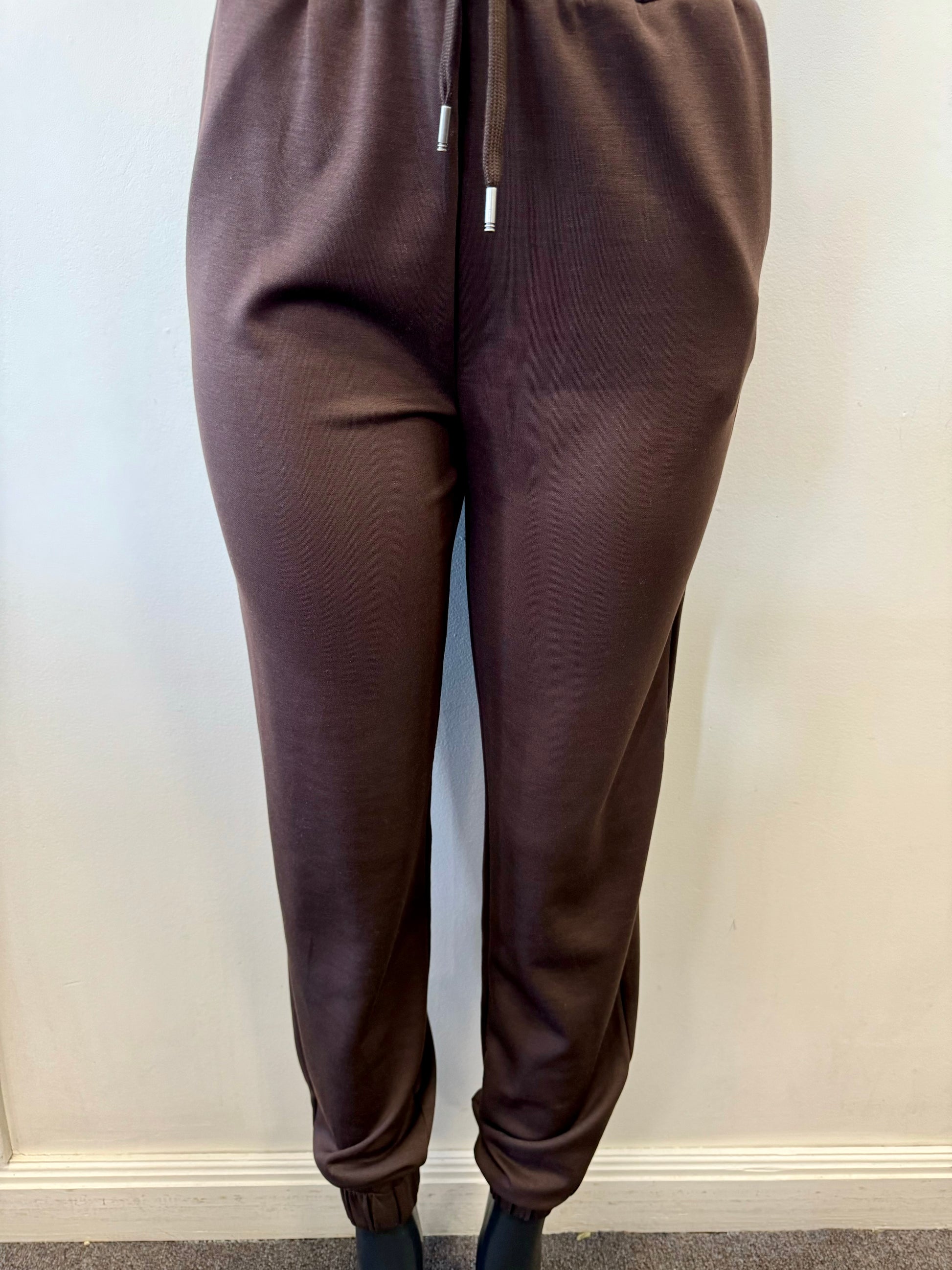 The image shows a person wearing SoyaConcept Banu 117 Relaxed Drawstring Pant in a brown jogger-style with a customizable fit, featuring a drawstring waist and elastic cuffs at the ankles, perfect for any casual occasion. They're standing against a plain light-colored wall.