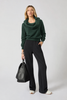 A woman with light hair stands against a plain background wearing the MPG Sport Dreamweave Off The Shoulder top in Scarab, styled with black wide-leg pants and white shoes, and carrying a large black handbag. The dark green, cowl-neck sweater is crafted from sustainable fabric.