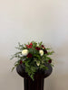 The Burgundy Christmas arrangement by Posies Flowers & Fashion features red and white carnations and chrysanthemums with green ferns, displayed on a dark round pedestal against a plain light-colored wall.