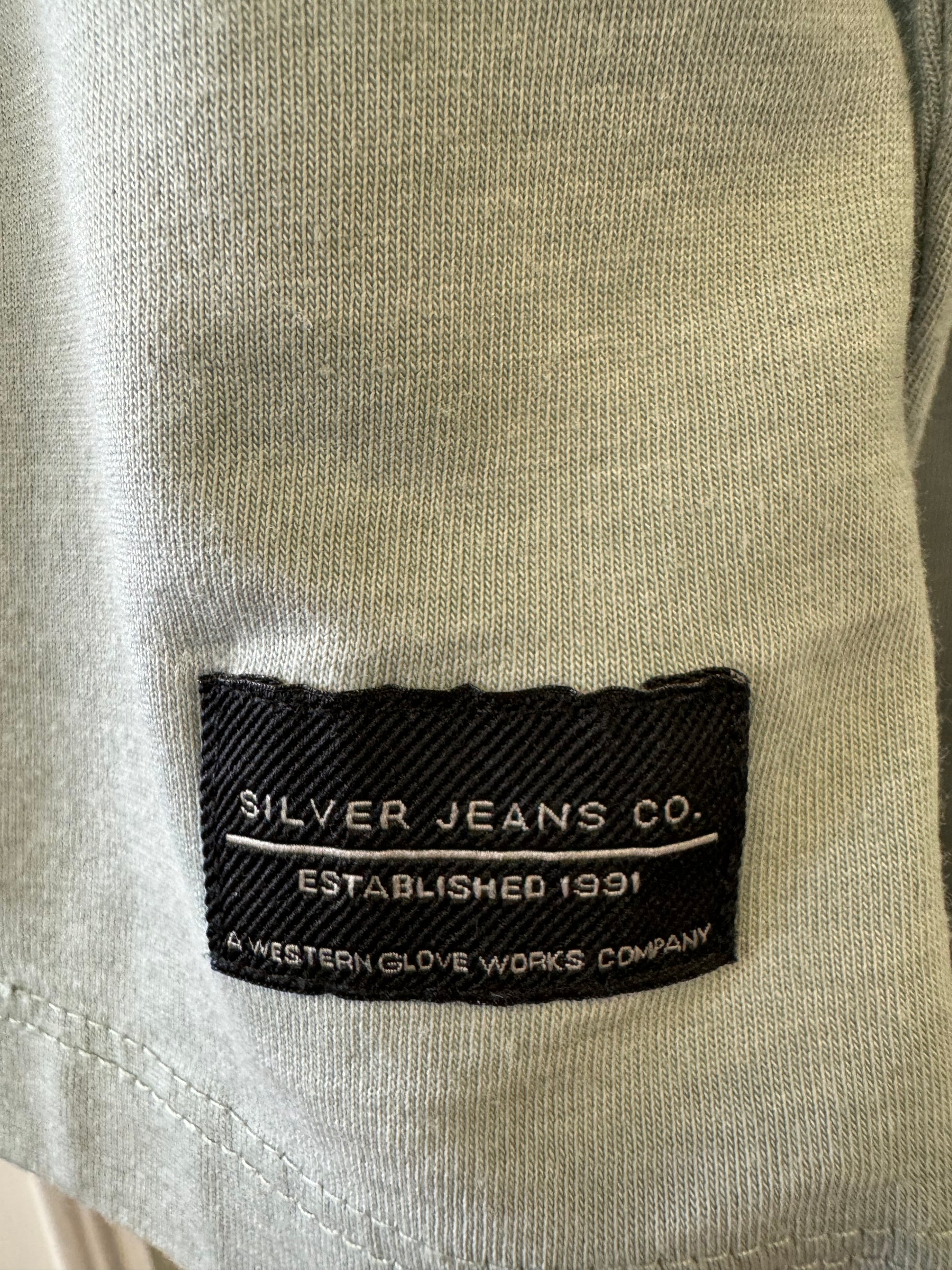 A mannequin dressed in the Silver Jeans Co. Shirt Long Sleeve - SAGE, featuring a classic 3-button design along the V-neckline. The sage green shirt, made from a comfortable cotton-polyester blend, is paired with black pants. The background is plain and white.