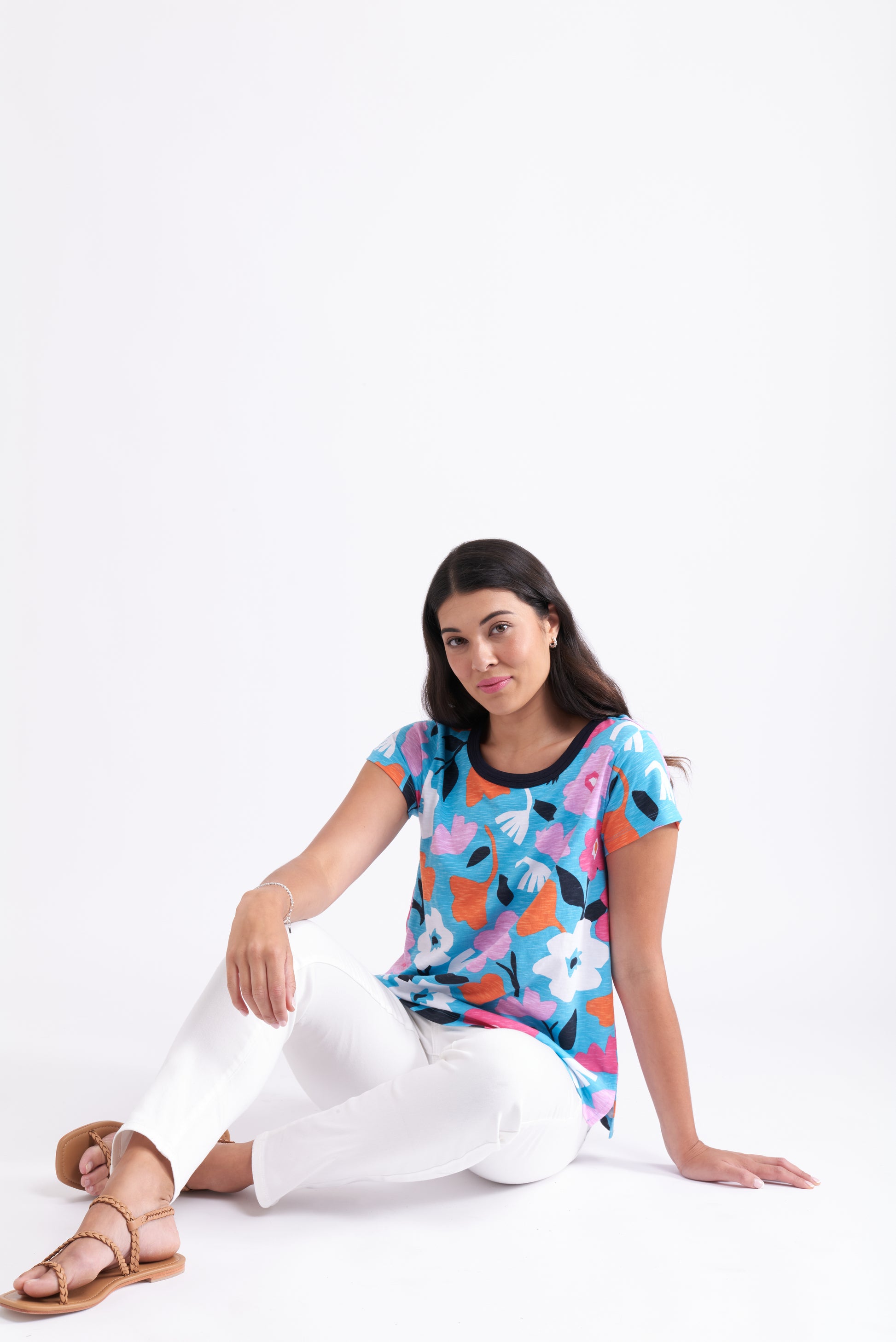 A woman with long dark hair sits against a white background, relaxed and wearing the FOIL Double Up Tee FO7495 Bold Bloom—a colorful floral 100% cotton top—paired with white pants and tan sandals.