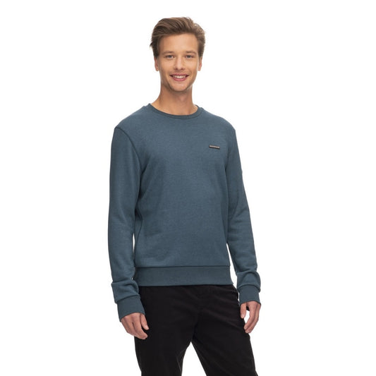 A smiling man with short brown hair is wearing a teal Ragwear Sweatshirt Pullover - INDDIE from RAGWEAR and black pants. He is standing against a plain white background, slightly turned to the side with his hands relaxed by his sides, showcasing a casual yet sporty look.