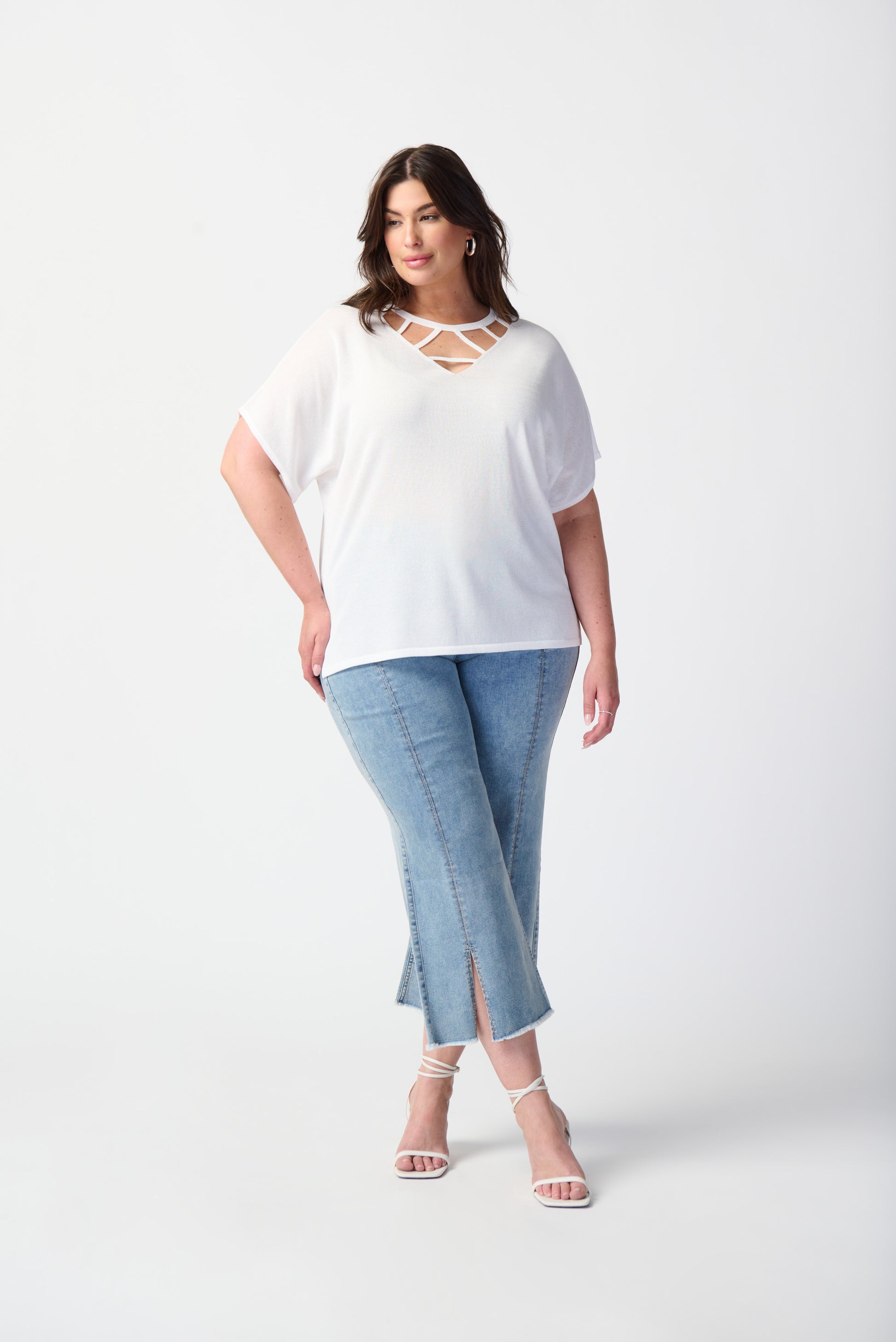 A woman stands on an outdoor balcony with greenery in the background. She is wearing a light knit sweater with a v-neck, 3/4 sleeves, and a split hem, paired with stylish Joseph Ribkoff's Culotte Jeans With Embellished Front Seam that have small slits at the bottom for a trendy edge. She has short hair and is accessorized with earrings.