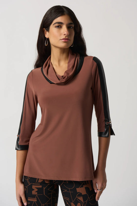 A woman with long dark hair is wearing the Joseph Ribkoff Toffee Faux-Leather Trim Tunic Style 233107, a brown long-sleeve top featuring black faux-leather trim on the shoulders and cuffs. She pairs it with black pants adorned with a geometric pattern. Standing against a plain white background, she gazes confidently at the camera.